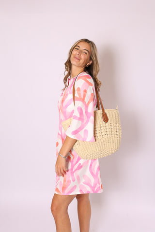 Woman wearing a pink and coral print dress, carrying the Sandy Shoulder Bag in cream straw with brown leather-look straps.