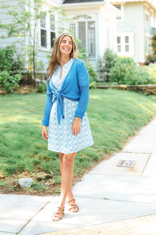Model wearing the Kara Knit Wrap in azure blue, tied over a patterned summer dress outdoors.