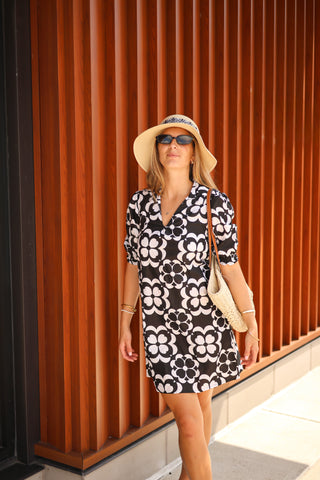 Lifestyle photo of woman in black and white floral Dune Mini Dress, styled with sun hat, sunglasses, and woven bag outdoors.