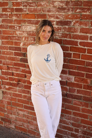 Woman wearing cream Seasonal Stitch Top with blue anchor embroidery, styled with white jeans, against a brick wall.