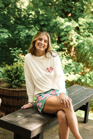 Woman sitting outdoors in cream Seasonal Stitch Top with red heart embroidery, styled with a green floral skirt.