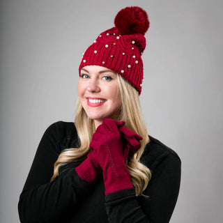Red knit hat with pom-pom decorated with pearls and gold balls, shown on model.